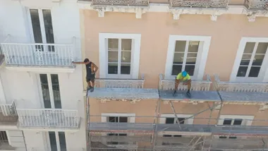 Dos albañiles encima de un andamio en un segundo piso sin arneses ni otras medidas de seguridad. Uno parace fumar un cigarro y el otro está asomado a la calle apoyado en un forjado.