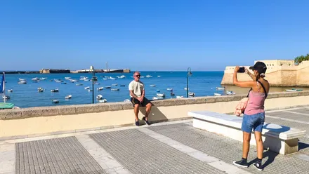 Una mujer le hace una foto a un hombre en la balaustrada La Caleta con el Castillo de San Sebastián de fondo en un día de cielo azulísimo