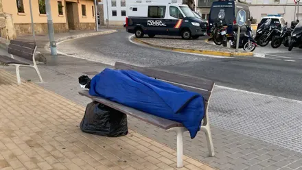 Una persona sin hogar duerme en un banco de la rotonda de Balbo en Cádiz, mientras al fondo se ve la rotonda engalanada con flores y decoración por el evento Cádiz Romano.