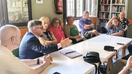 Imagen de una mesa de reunión en Cádiz. A la izquierda, Óscar Torres y José Ramón Ortega, representantes del PSOE, conversan con Flor Muro y otros miembros de la junta directiva de la Asociación de Vecinos de Muñoz Arenillas. Todos están sentados, con papeles y documentos sobre la mesa, en un ambiente de trabajo y diálogo.