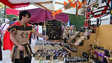 Hombre vestido con casco y coraza de legionario romano coloca productos sobre una mesa de madera decorada con telas rojas.