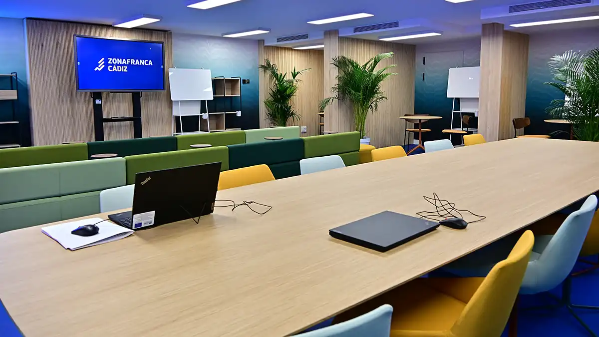 Sala rectangular con mesa central alargada, sillas a su alrededor y una pantalla interactiva al fondo. Un grupo pequeño de personas conversa durante la visita institucional.
