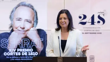 Patricia Cavada, con chaqueta blanca presentando los actos del 24-S, tras ella una foto de Joan Manuel Serrat.