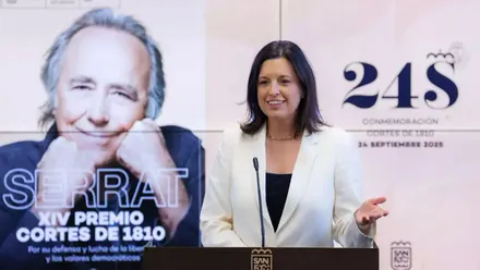 Patricia Cavada, con chaqueta blanca presentando los actos del 24-S, tras ella una foto de Joan Manuel Serrat.