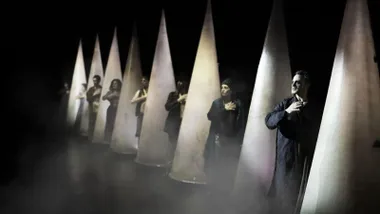 Intérpretes de una obra de teatro en agarrados a conos blancos, predominio de la oscuridad.