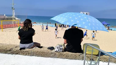 Una pareja observa un partido de fútbol plata en La Victoria mientras el hombre se protege con una sombrilla apoyada en su hombro.