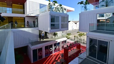 Imagen panorámica del complejo Incubazul, formado por contenedores marítimos reciclados apilados en varios niveles. Se aprecian sus terrazas abiertas, la fachada modular y el entorno urbano de la Zona Franca de Cádiz, con visitantes recorriendo el acceso principal.