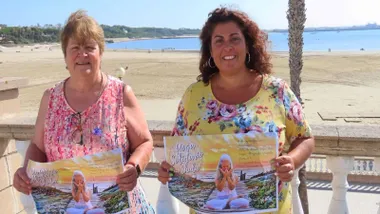 Delegada de Cultura de Rota y organizadora sosteniendo el cartel en la mano y tras ellas la playa.