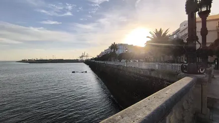 Vista de la bahía de Cádiz en una mañana despejada, con el sol brillando detrás de un edificio y las grúas del muelle recortadas en el horizonte, tomadas desde la Alameda.
