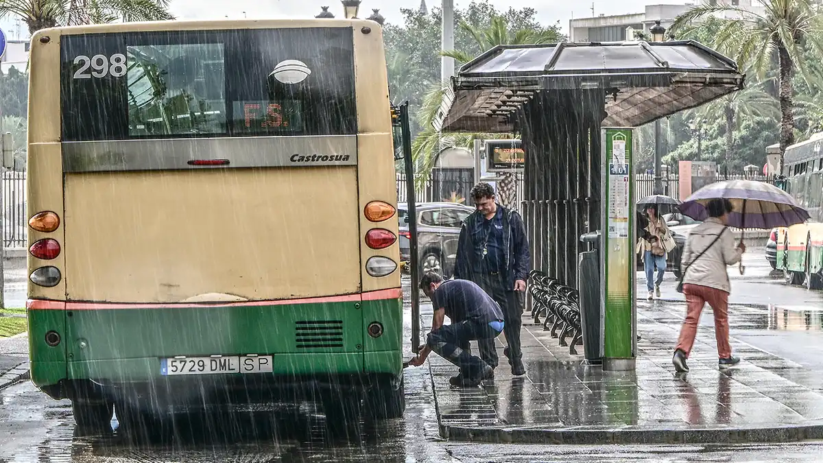 Dos operarios intentan reparar un autobús detenido en la Plaza de España de Cádiz. Llueve con fuerza, el pavimento está mojado y varios peatones pasan con paraguas mientras otro autobús espera a unos metros.