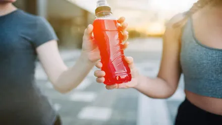 Dos chicas corriendo y de pasan, con la mano, una botella de bebida isotónica naranja.