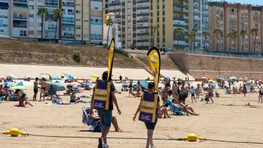 Educadores ambientales de Cada Lata Cuenta, con mochilas amarillas recolectoras, entre bañistas en la playa de Santa María del Mar en Cádiz durante una campaña de reciclaje de latas.