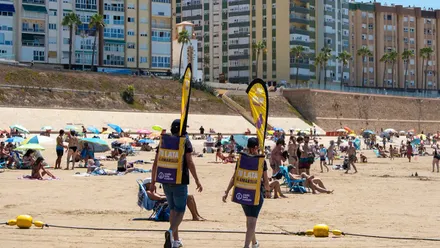 Educadores ambientales de Cada Lata Cuenta, con mochilas amarillas recolectoras, entre bañistas en la playa de Santa María del Mar en Cádiz durante una campaña de reciclaje de latas.