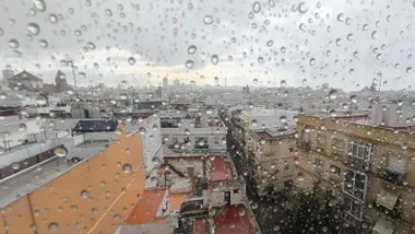 Primer plano de una ventana con gotas de lluvia en Cádiz; al fondo, la catedral y edificios del centro bajo un cielo gris.
