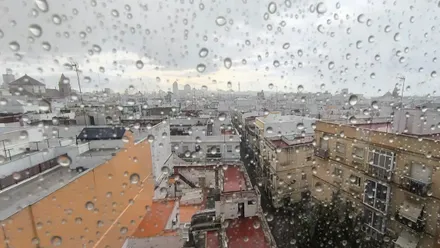 Primer plano de una ventana con gotas de lluvia en Cádiz; al fondo, la catedral y edificios del centro bajo un cielo gris.