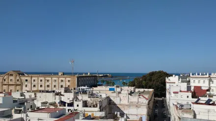 Vista luminosa de la playa de La Caleta en Cádiz bajo un cielo despejado. En primer plano se distingue el edificio histórico de Valcárcel y, al fondo, el Castillo de San Sebastián.