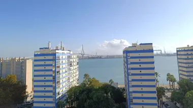 Vista de la barriada de la Paz en Cádiz bajo un cielo completamente despejado. Al fondo se distingue el segundo puente sobre la bahía, con luz clara y algunas nubes.