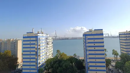 Vista de la barriada de la Paz en Cádiz bajo un cielo completamente despejado. Al fondo se distingue el segundo puente sobre la bahía, con luz clara y algunas nubes.