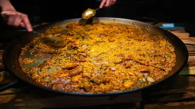Unas manos con una cuchara sirven paella de la paellera enorme. Destaca el arroz amarillo típico.