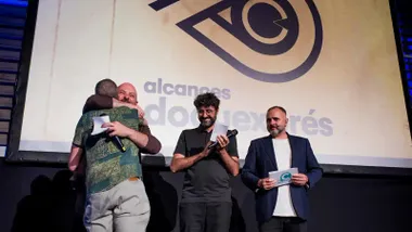 El director Lolo Ruiz se abraza al jurado al subir al escenario tras ganar el premio DocuExprés en el Festival Alcances 2025, con el logo del festival proyectado en pantalla de fondo.