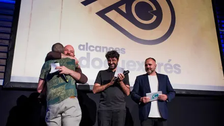 El director Lolo Ruiz se abraza al jurado al subir al escenario tras ganar el premio DocuExprés en el Festival Alcances 2025, con el logo del festival proyectado en pantalla de fondo.