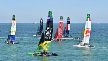 Catamarán de Gran Bretaña navegando frente a la costa de Cádiz durante la final de la SailGP 2025, adelantando al barco de Nueva Zelanda mientras miles de espectadores siguen la regata desde el Paseo de Santa Bárbara.