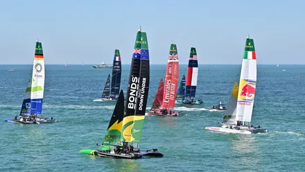 Catamarán de Gran Bretaña navegando frente a la costa de Cádiz durante la final de la SailGP 2025, adelantando al barco de Nueva Zelanda mientras miles de espectadores siguen la regata desde el Paseo de Santa Bárbara.