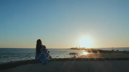 Mujer de espaldas frente al mar en la playa de La Caleta de Cádiz, con el castillo de San Sebastián al fondo y el sol poniéndose en el horizonte.