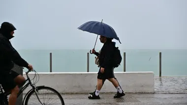 Dos personas caminan bajo la lluvia en el paseo Marñitimo sobre Santa Maria del Mar con un paraguas y otra se protege del agua mientras va en su bicicleta.
