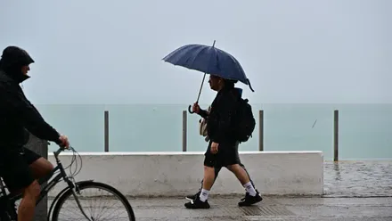 Dos personas caminan bajo la lluvia en el paseo Marñitimo sobre Santa Maria del Mar con un paraguas y otra se protege del agua mientras va en su bicicleta.