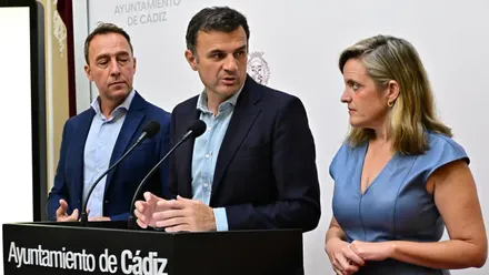 El alcalde de Cádiz, Bruno García, comparece junto a José Manuel Cossi y Mayte González ante un estrado con dos micrófonos. Detrás de ellos se distinguen el logo institucional del Ayuntamiento de Cádiz y el escudo municipal sobre un fondo blanco.