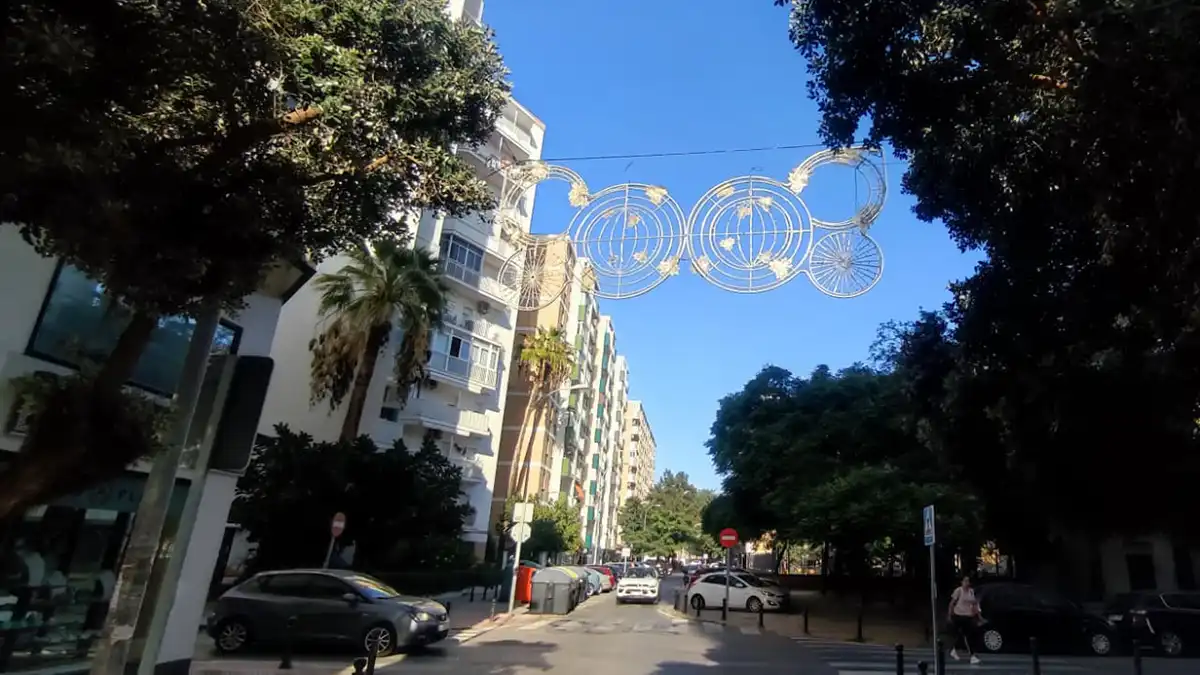 Fotografía del barrio de La Laguna en Cádiz con el alumbrado navideño instalado. Se ven estructuras de luces con formas geométricas y colores dorados suspendidas sobre la calle, con árboles y viviendas y la Plaza Reina Sofía y una iglesia.