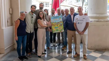 En la imagen, varias personas posan en el balcón del Ayuntamiento de Cádiz sosteniendo el cartel de la III Milla Popular. Entre ellas se encuentran la concejala Gloria Bazán, el edil de Deportes Carlos Lucero y miembros de la Asociación de Vecinos Murallas de San Carlos y los Scouts de Cádiz.
