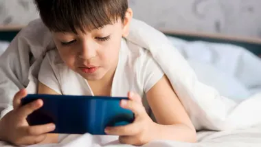 Niño pequeño en su cama, tapado con la sábana, usando un móvil.