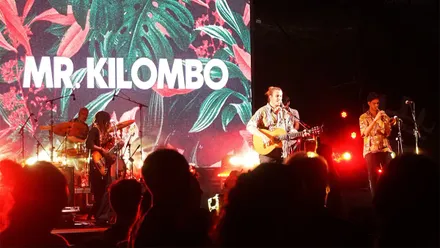 El cantante Mr. Kilombo aparece en el escenario con su guitarra, iluminado por focos de colores, mientras interpreta una canción frente al público durante un concierto.
