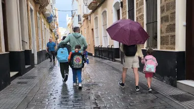 Un padre acompaña a una niña al colegio con un paraguas mientras otro lo hace con dos niños sin usarlo.