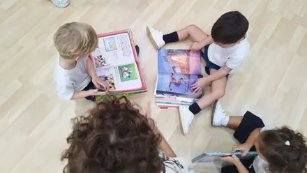 Dos niños sentados en el suelo de una biblioteca municipal de Cádiz hojean libros infantiles, rodeados de estanterías con cuentos y luz natural. Imagen que representa la lectura en un entorno público y accesible para todos.