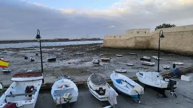 Vista de la playa de La Caleta en Cádiz con la marea baja y varias barcas varadas en primer plano; al fondo se distinguen los castillos de Santa Catalina y San Sebastián bajo un cielo gris y nublado.