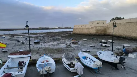 Vista de la playa de La Caleta en Cádiz con la marea baja y varias barcas varadas en primer plano; al fondo se distinguen los castillos de Santa Catalina y San Sebastián bajo un cielo gris y nublado.