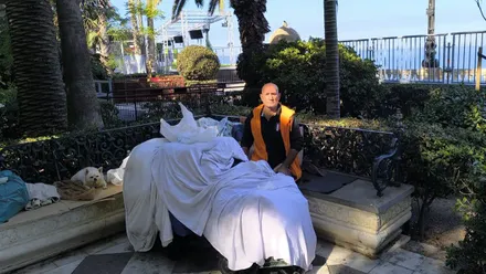 Hombre sin hogar sentado en un banco con sus pertenencias en los Jardines de Carlos III; detrás se levantan las gradas y el césped artificial de la SailGP en Cádiz.
