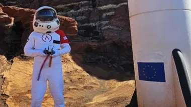 Hombre vestido de astronauta y cohete a su lado.