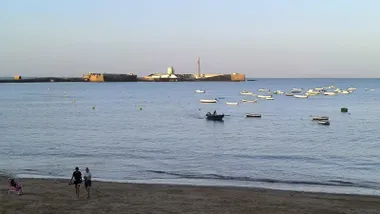 Una pequeña embarcación navegando a remos entre otras ancladas en la playa de La Caleta, en Cádiz, con el Castillo de San Sebastián al fondo y dos jóvenes haciendo ejercicio en la orilla bajo un cielo despejado.