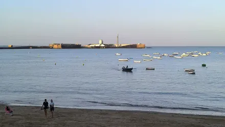 Una pequeña embarcación navegando a remos entre otras ancladas en la playa de La Caleta, en Cádiz, con el Castillo de San Sebastián al fondo y dos jóvenes haciendo ejercicio en la orilla bajo un cielo despejado.