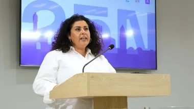 Laura Almisas con camisa blanca y fondo con una pantalla en la que se lee "25N".