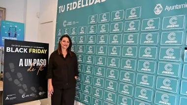 Imagen de Yolanda Pérez, gerente de Cádiz Centro Comercial Abierto, sonriendo mientras posa de pie junto a un cartel vertical que muestra los lemas de las campañas “Tu fidelidad abre regalos” y “Black Friday. A un paso de ti”. Detrás se distingue un fondo luminoso con colores otoñales y el logotipo del CCCA