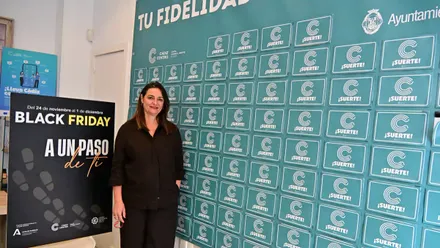 Imagen de Yolanda Pérez, gerente de Cádiz Centro Comercial Abierto, sonriendo mientras posa de pie junto a un cartel vertical que muestra los lemas de las campañas “Tu fidelidad abre regalos” y “Black Friday. A un paso de ti”. Detrás se distingue un fondo luminoso con colores otoñales y el logotipo del CCCA