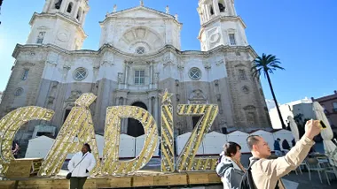 Una mujer posa delante de un acrtel de Cádiz de la iluminación de Navidad delante de la Catedral de Cádiz mientras otro turista se hace un selfie con las letras de fondo.