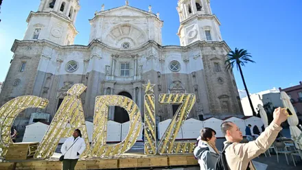 Una mujer posa delante de un acrtel de Cádiz de la iluminación de Navidad delante de la Catedral de Cádiz mientras otro turista se hace un selfie con las letras de fondo.