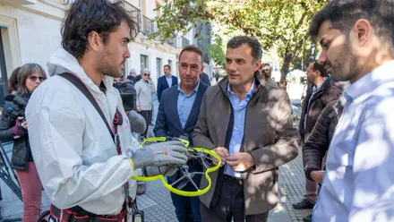 En la imagen se ve al presidente de Aguas de Cádiz, José Manuel Cossi, de pie junto al alcalde de Cádiz en un espacio abierto. Ambos observan el equipo técnico preparado para volar un pequeño dron utilizado para inspeccionar tuberías y colectores. El dron aparece en primer plano sobre el suelo, rodeado de personal técnico y material de trabajo.