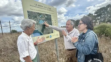 Fotografía de Esther Gil de Reboleño junto a representantes de colectivos ecologistas, reunidos frente a un cartel informativo de la Laguna de La Janda. La imagen muestra a la diputada conversando con los activistas mientras señalan detalles del panel, en un entorno exterior y con luz natural. El cartel del humedal aparece claramente visible como elemento central de la escena.